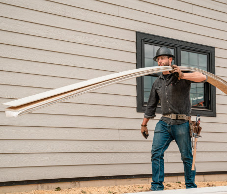 A worker carrying several lengths of siding across his shoulder, walking past the side of a house.